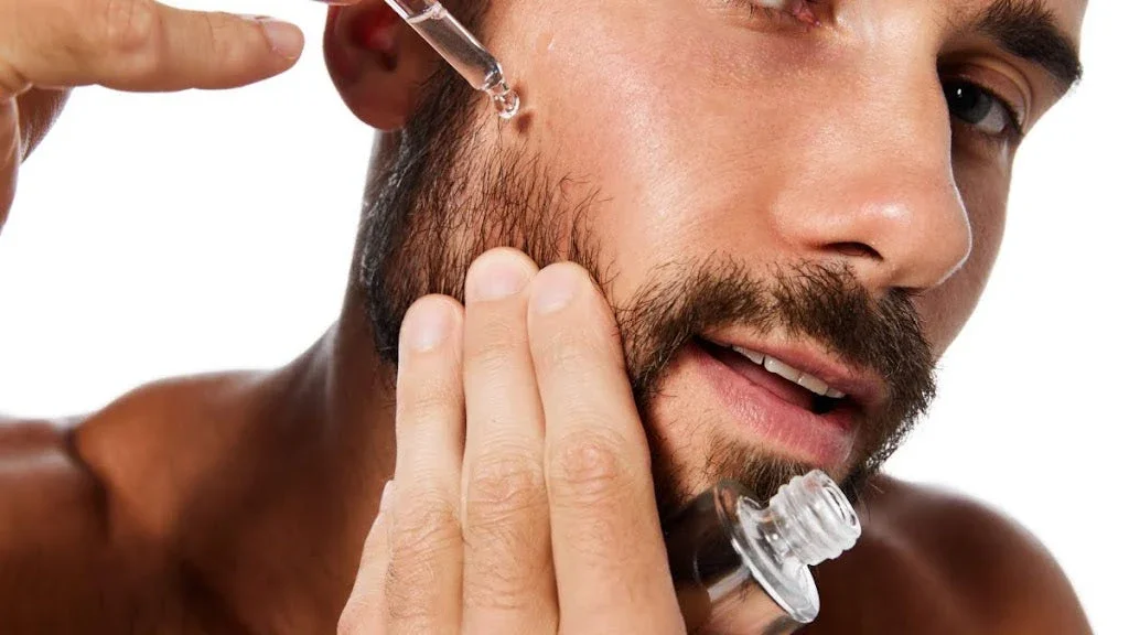 Close-up of a bare-chested man pointing towards his ear, which has a visible earring. The background is plain white.